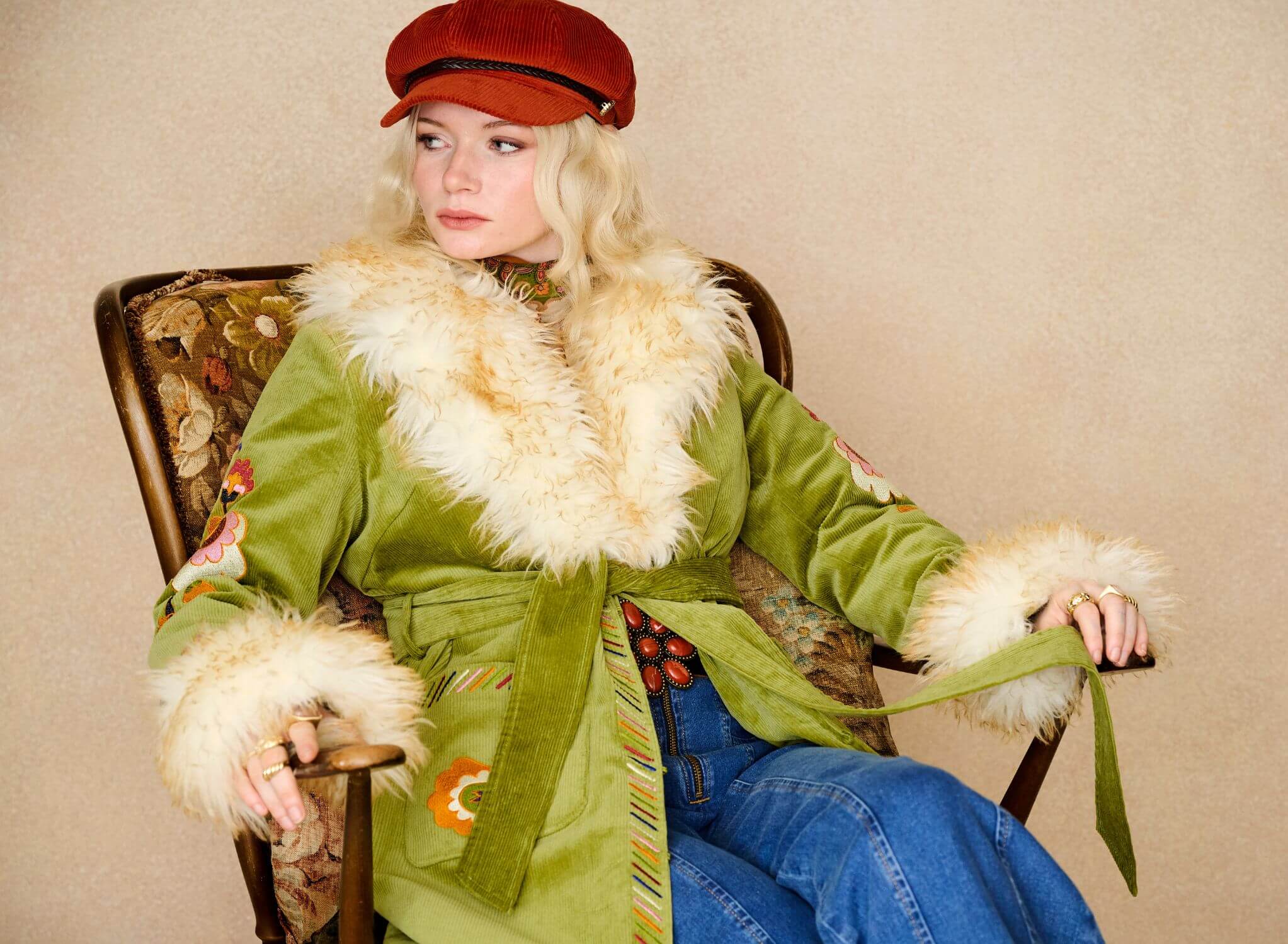 Model sitting down wearing The Hippie Shake green embroidered Penny Lane coat with faux fur trim and blue denim jeans.