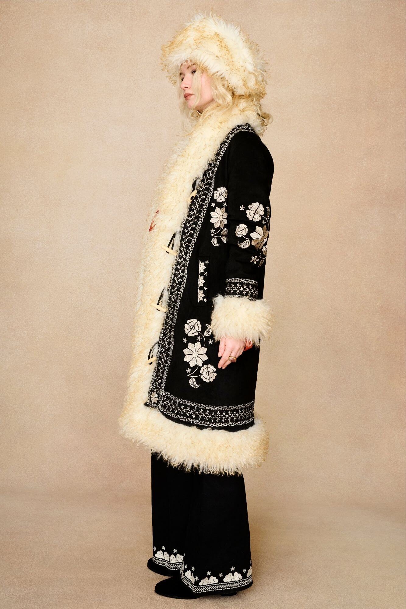 Model wears cream faux fur hat with vintage detailing and embroidered finish. A 70s-inspired boho winter hat made for statement styling by The Hippie Shake - side view with Black Penny Lane Coat