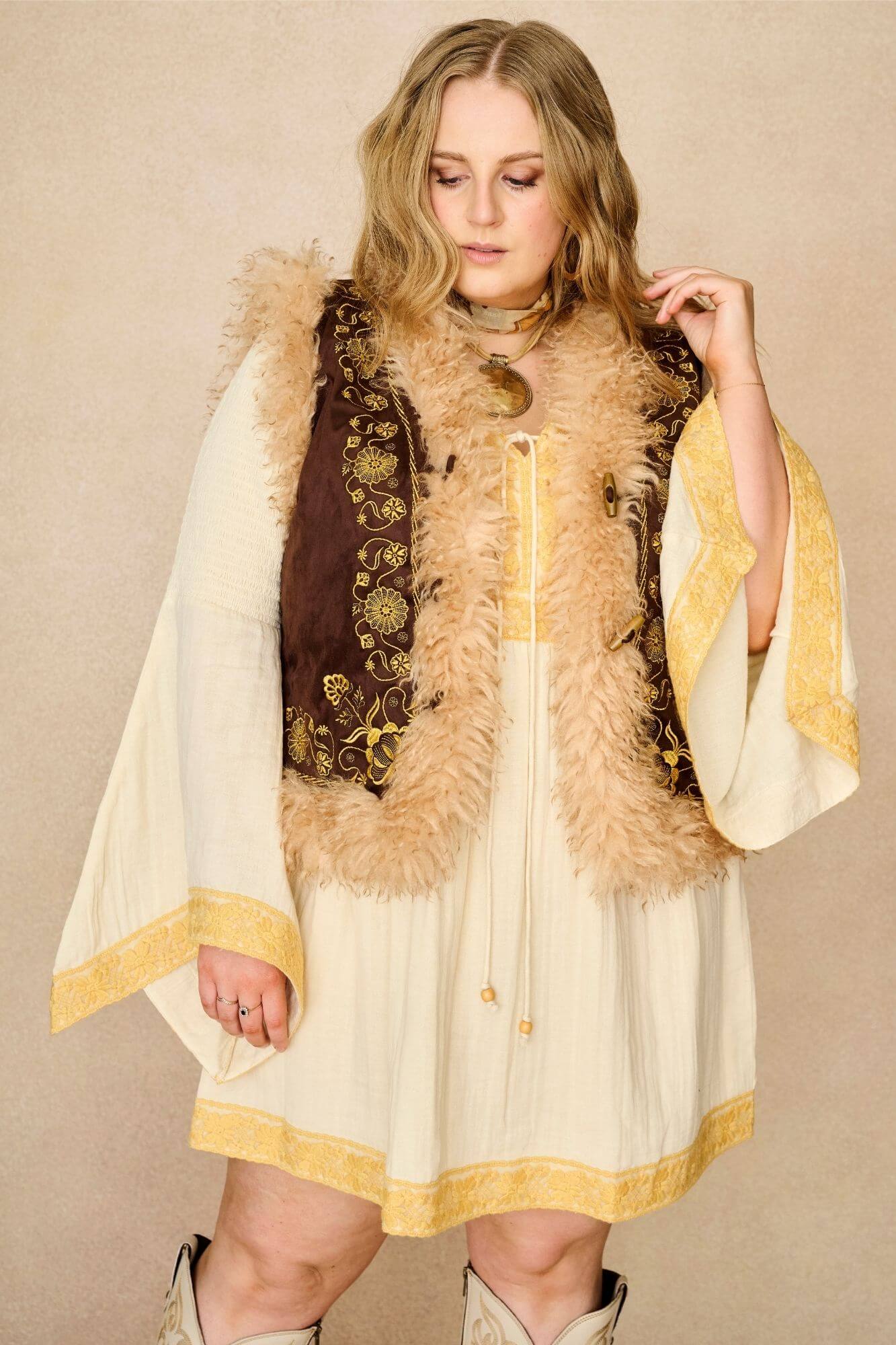 Model wears brown embroidered Penny Lane vest in faux suede with faux fur trim. A 70s-inspired boho layering piece made for vintage and festival styling by The Hippie Shake - front view paired with cream dress