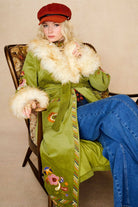 Model wears green embroidered Penny Lane coat in cotton corduroy with faux fur trim. A 70s-inspired statement coat made for bohemian, vintage styling by The Hippie Shake - sitting down view