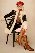 Model wears dark brown embroidered Penny Lane coat in faux suede with faux fur trim by The Hippie Shake. A 70s-inspired statement coat made for bold, vintage boho styling - sitting down