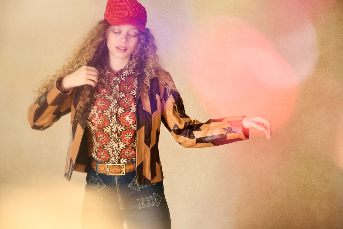 Model wears a retro printed blouse under a 70s inspried patchwork leather jacket by The Hippie Shake and a red baker hat and jeans