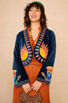 Model wearing The Hippie Shake blue velvet embroidered jacket with Zodiac symbols and orange dress against a beige background