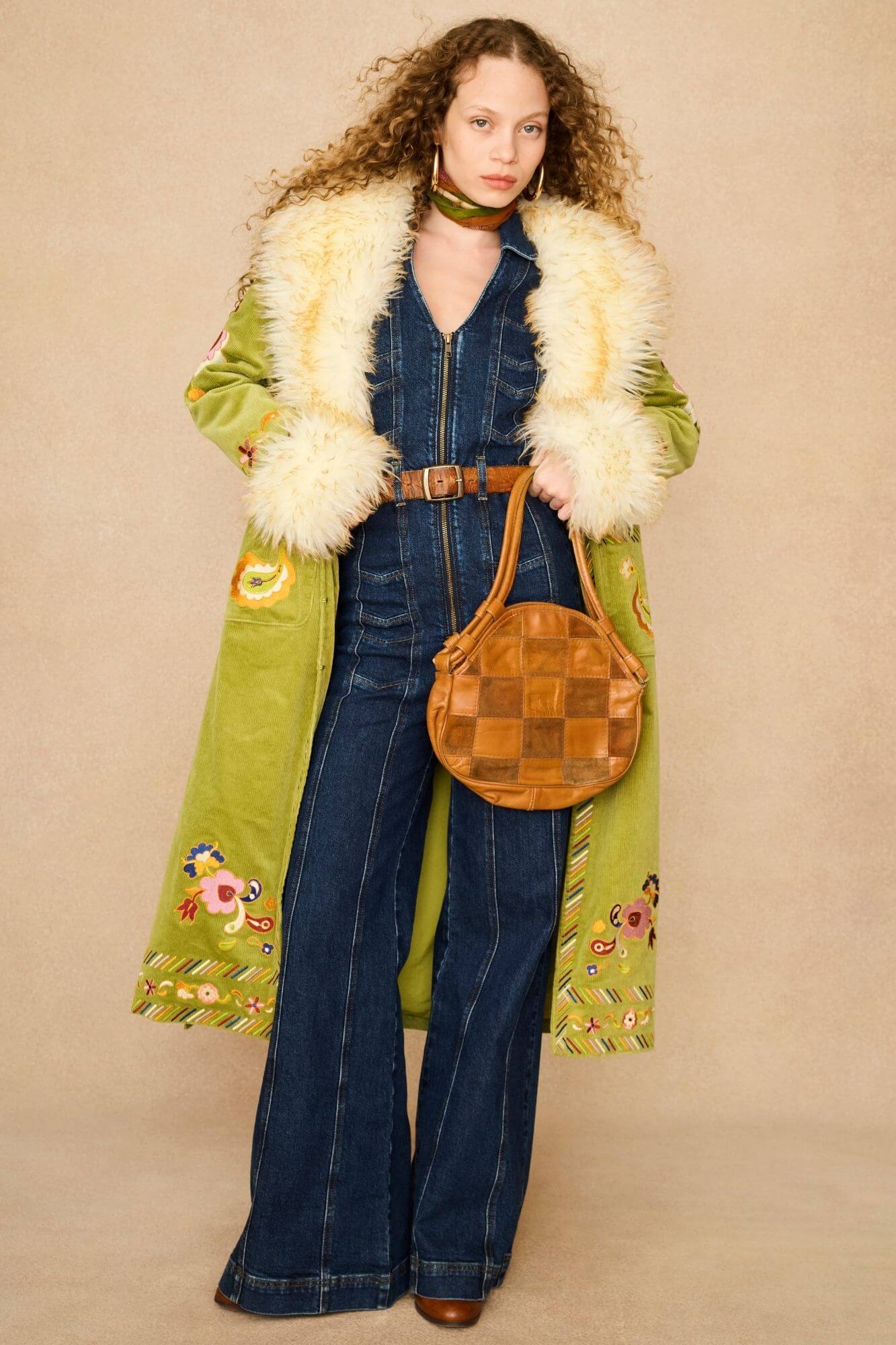 A model wearing a long olive green coat with a cream faux fur collar, a dark blue denim jumpsuit, and a brown woven round handbag, front view.