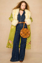 A model wearing a long olive green coat with a cream faux fur collar, a dark blue denim jumpsuit, and a brown woven round handbag, front view.