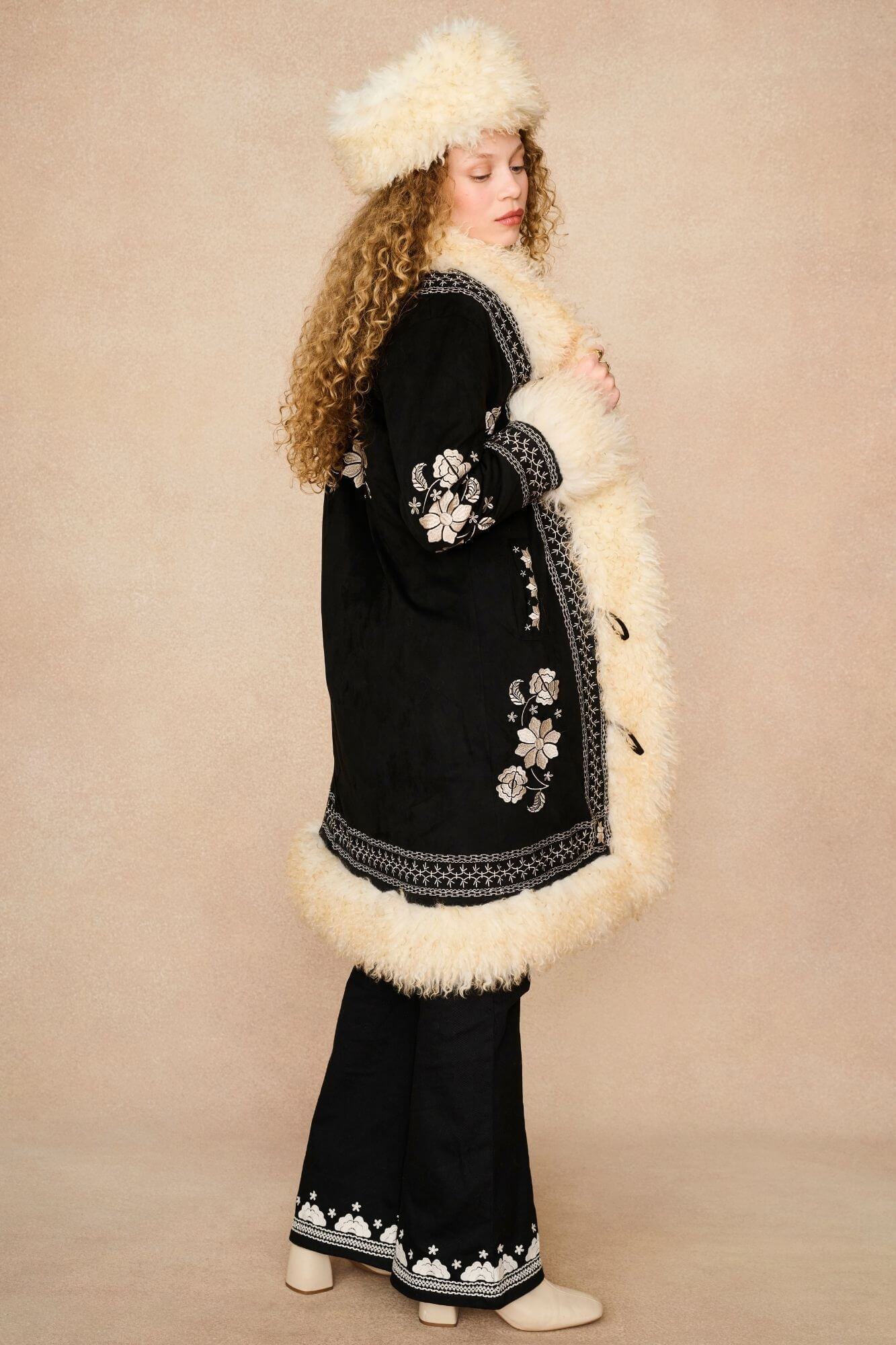 A model wearing a black 1970s boho coat with cream faux fur trim, a matching cream fur hat, and black flared pants by the Hippie Shake