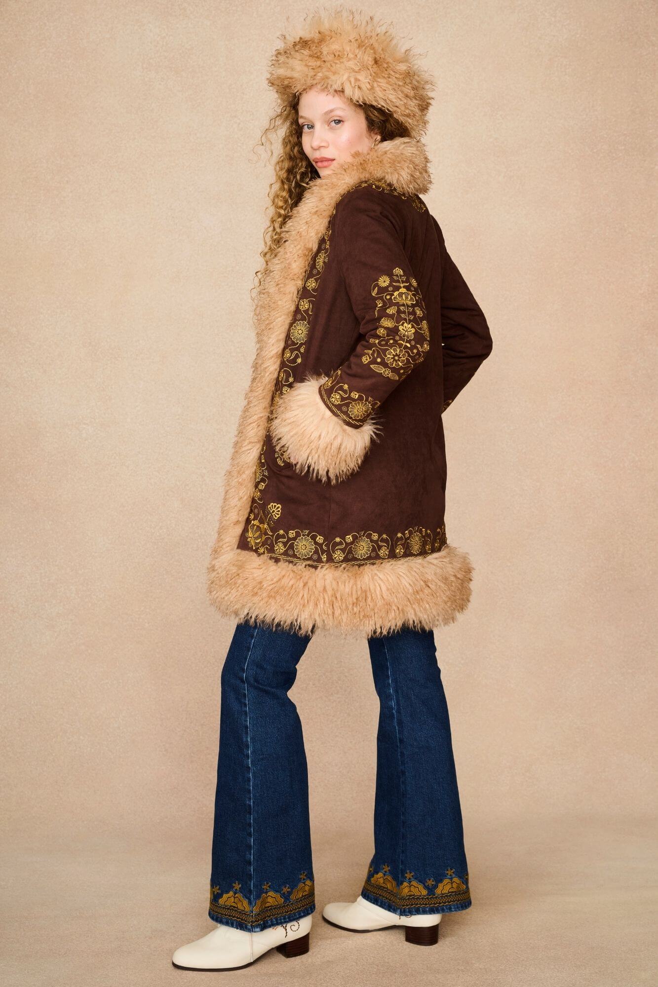 A model wearing a 70s bohemian-style outfit featuring a brown penny lane jacket with beige fur trim and a matching fur-trimmed hat.