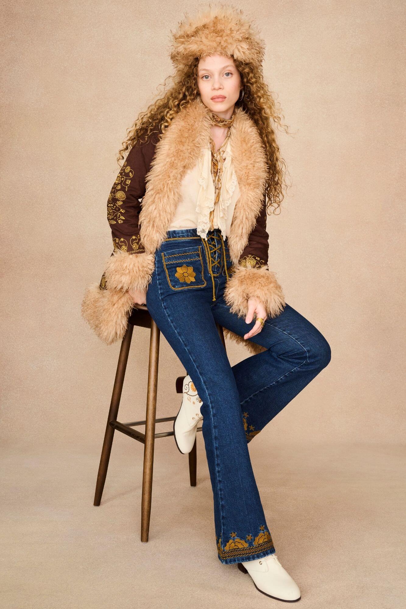 A model wearing a 70s bohemian-style outfit featuring a brown penny lane jacket with beige fur trim and a matching fur-trimmed hat.
