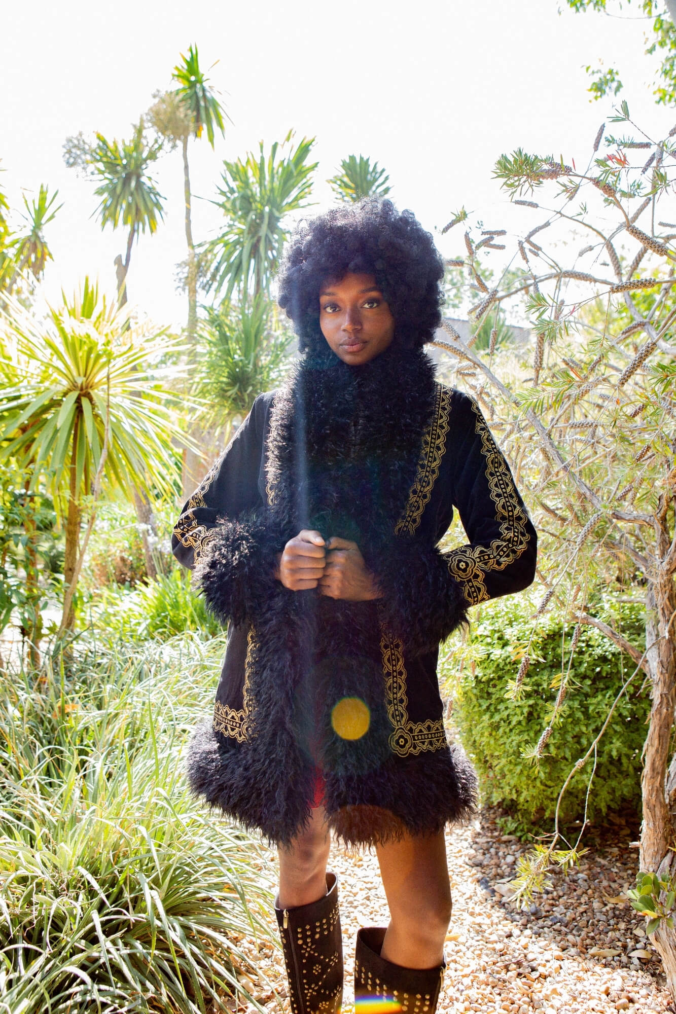 A model wearing a black fringed jacket with gold embroidered details, a matching fringed scarf, and black studded boots, standing in a lush green outdoor garden setting.