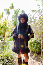 A model wearing a black fringed jacket with gold embroidered details, a matching fringed scarf, and black studded boots, standing in a lush green outdoor garden setting.