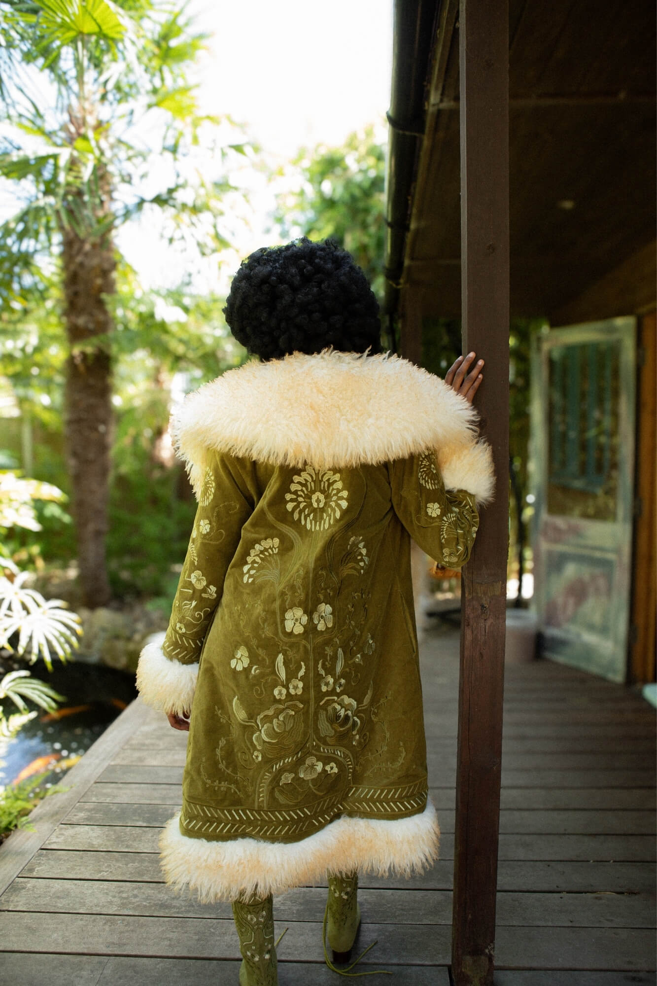 Model viewed from behind wearing a long olive green Penny Lane embroidered coat with a large cream fur collar and hem, standing on a wooden deck outdoors near palm trees and a porch.