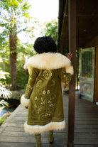 Model viewed from behind wearing a long olive green Penny Lane embroidered coat with a large cream fur collar and hem, standing on a wooden deck outdoors near palm trees and a porch.