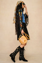 A woman models a black embroidered bohemian-style dress with a blue shirt underneath, a patterned headscarf, black lace-up knee-high boots, and a round tan leather shoulder bag, set against a solid beige studio background.
