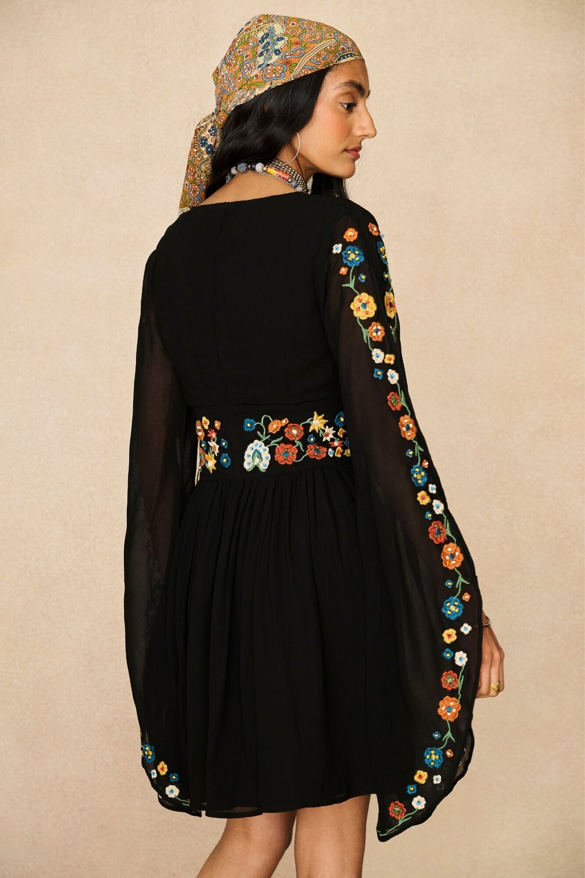 A woman shown from the back wearing a black dress with colorful floral embroidery along the waist and sleeves, paired with a patterned headscarf, against a plain beige background.