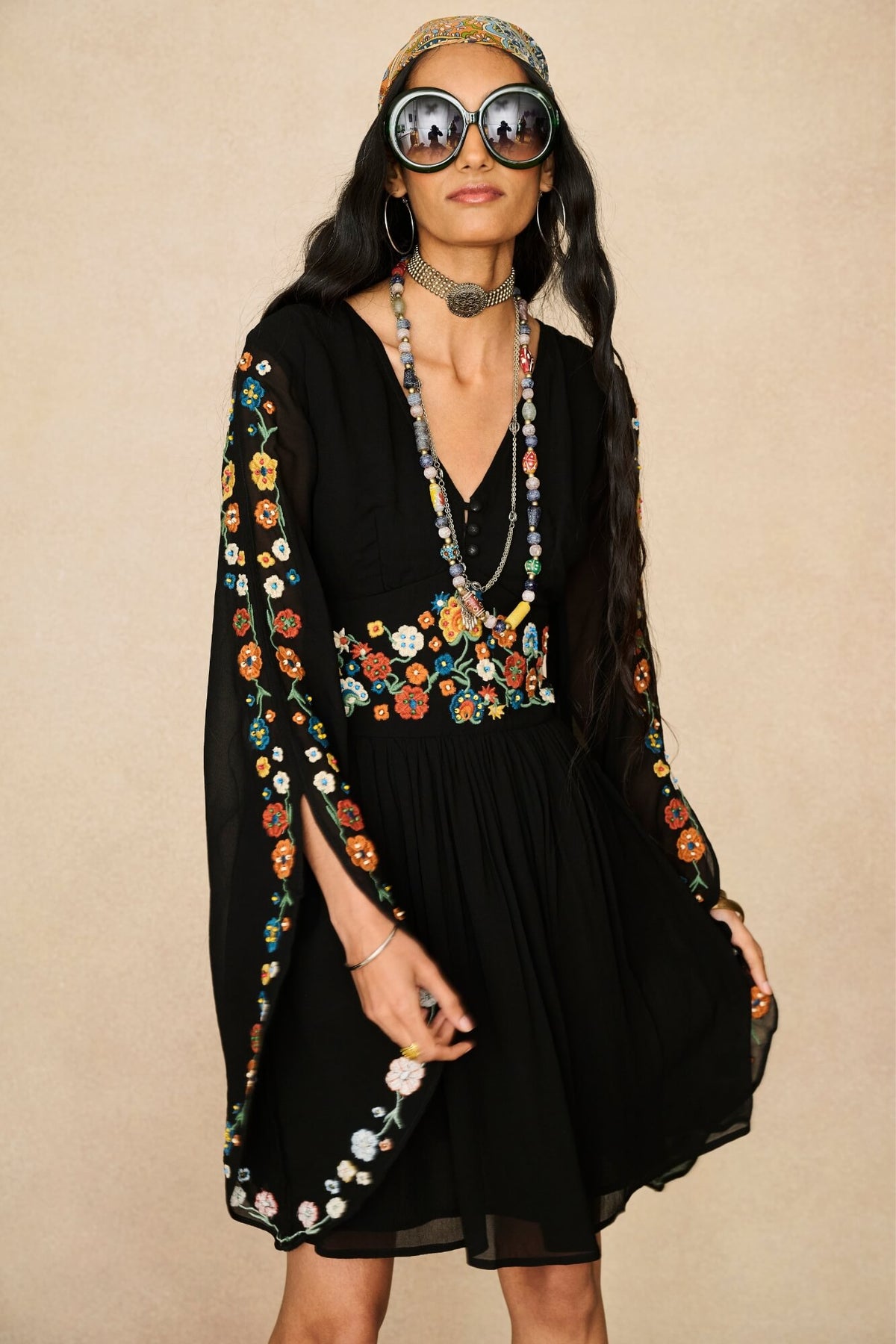 A woman modeling a black bohemian-style dress with colorful floral embroidery on the sleeves and waist, accessorized with large round sunglasses, a patterned headband, and layered necklaces against a beige background.