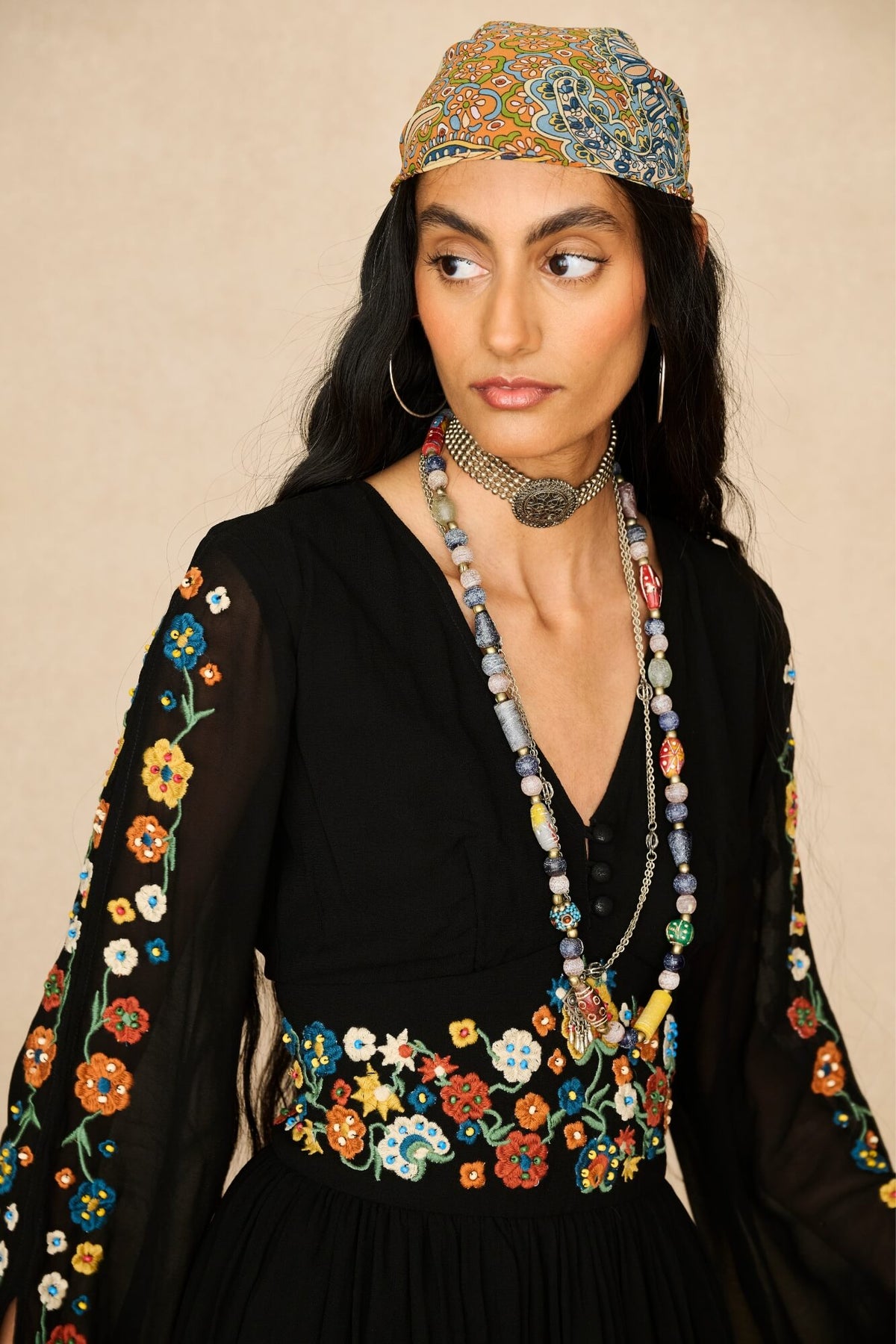 A woman wearing a black embroidered dress with colorful floral patterns on the sleeves and waist, accessorized with layered beaded necklaces and a patterned headscarf, against a plain beige background.