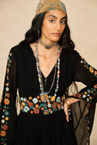 A woman wearing a black embroidered dress with colorful floral patterns on the sleeves and waist, accessorized with layered necklaces, a headpiece, and gold jewelry, set against a plain beige background.