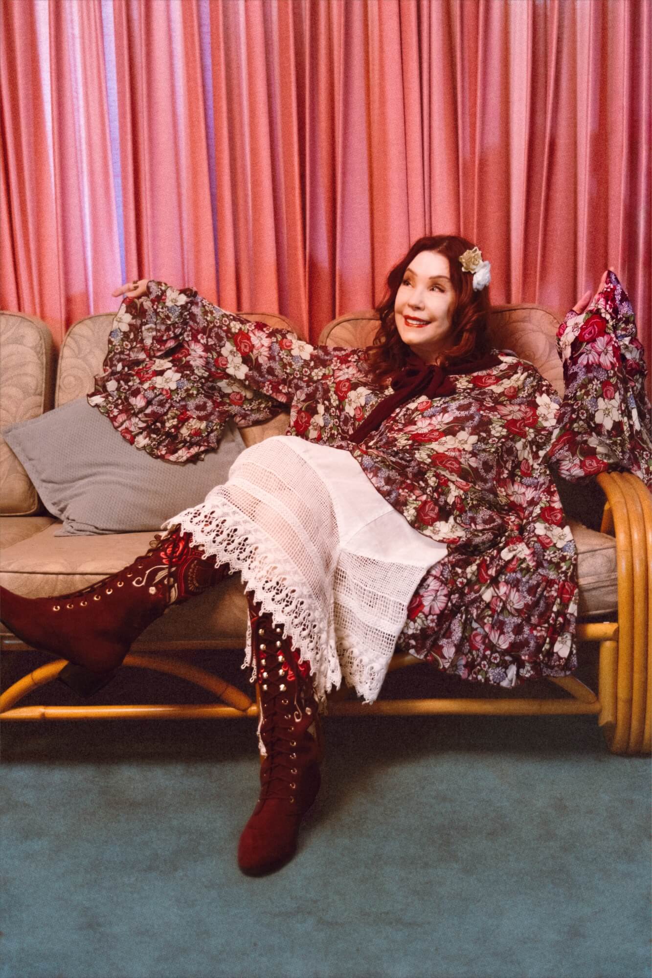 Woman in a floral top sitting on a couch with pink curtains in the background