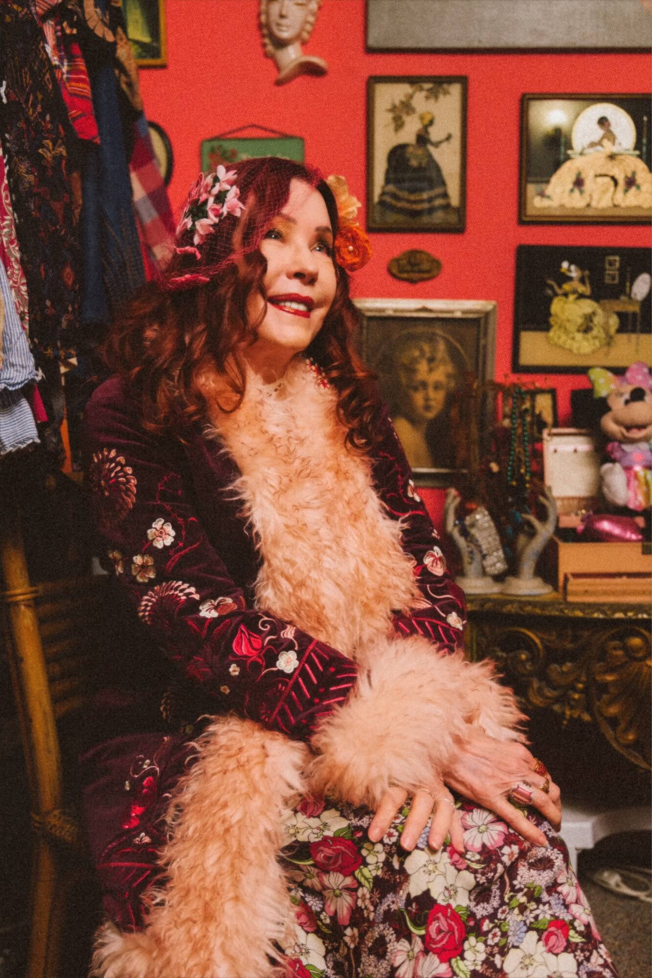 Woman in a floral dress with a fur coat sitting in a room with red walls and various decorations.