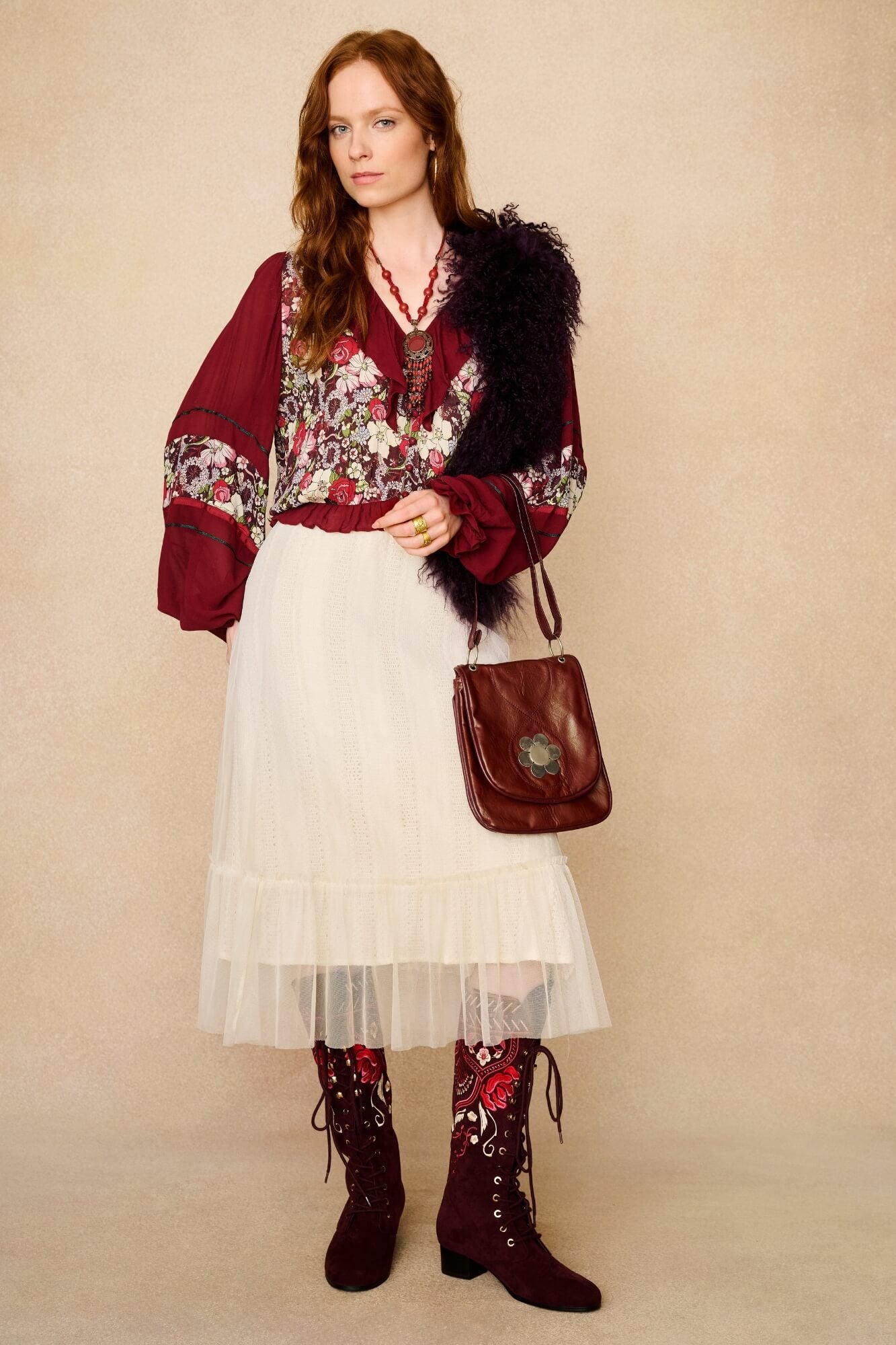 Woman wearing a floral blouse, red cardigan, white skirt, and brown boots with a brown handbag against a beige background.