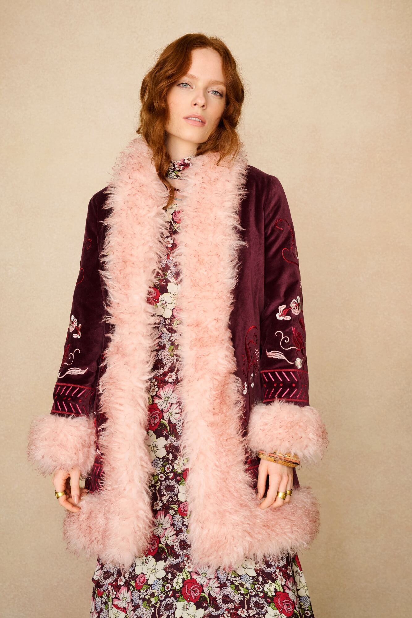 Woman wearing a floral dress with a burgundy jacket and pink feathered scarf against a beige background