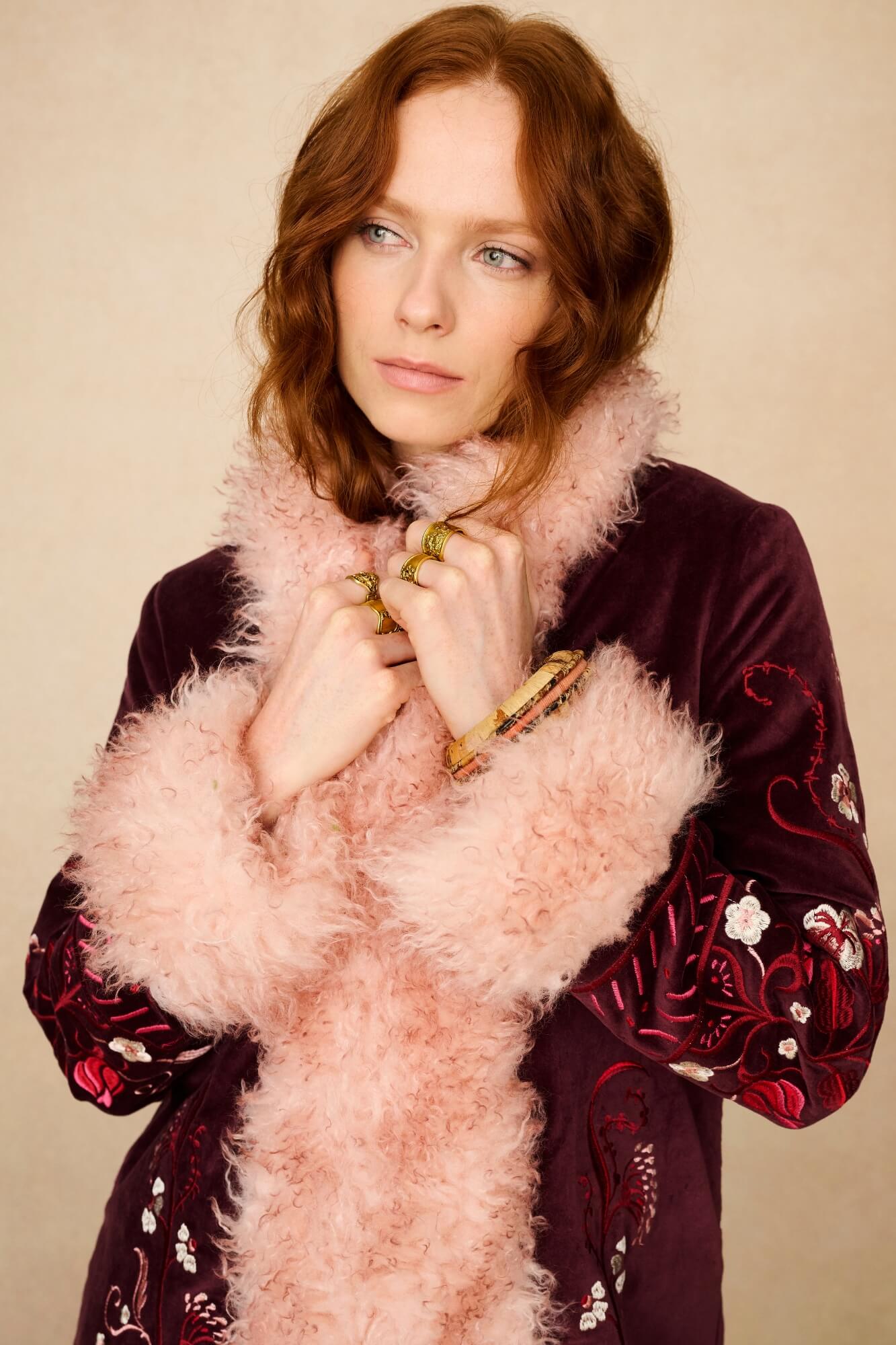 Woman wearing a burgundy embroidered dress with a pink fluffy scarf against a beige background - close up