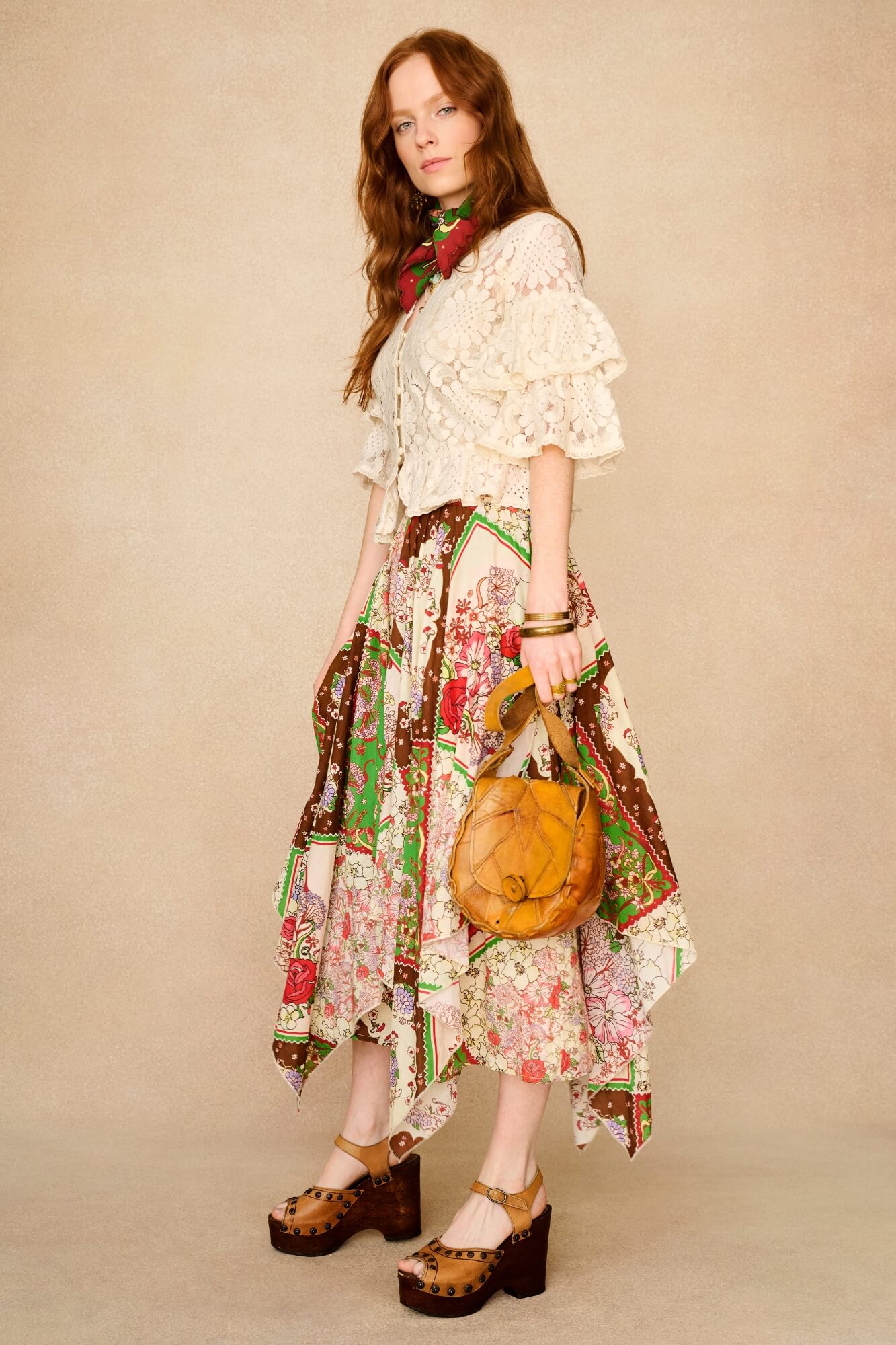 Woman wearing a floral dress with a patterned top, holding a brown leather handbag against a beige background.