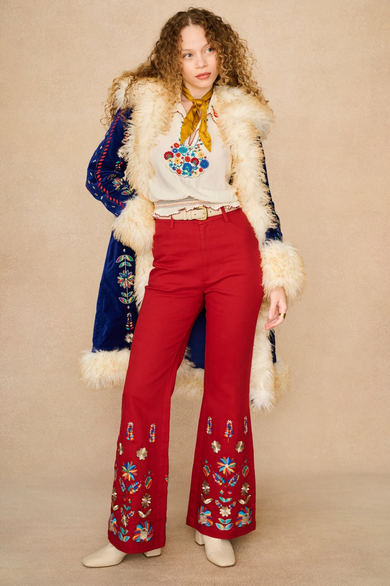 A model stands against a beige studio backdrop wearing a retro-style outfit featuring bright red flared pants with colorful floral embroidery near the hem, a cream blouse with a multicolor floral chest print, a mustard yellow neck scarf, and a blue velvet jacket with white faux fur trim.
