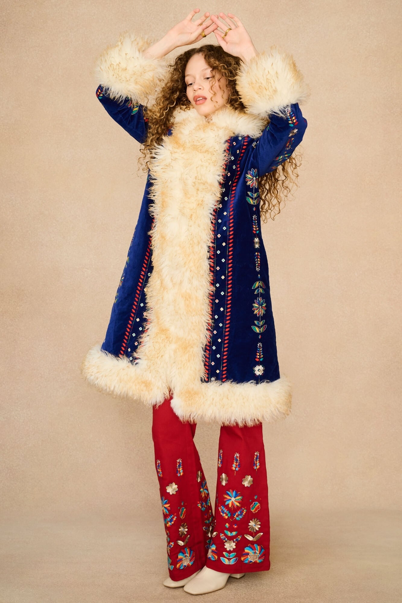 Model in a vintage-style bohemian Penny Lane Coat by The Hippie Shake with cream faux-fur trim, blue embroidered panels, and red embroidered pants against a solid beige background.