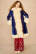 A model wearing a long blue velvet Penny Lane coat with a thick cream fur collar and cuffs, paired with red wide-leg pants featuring colorful embroidery, set against a plain beige studio background.