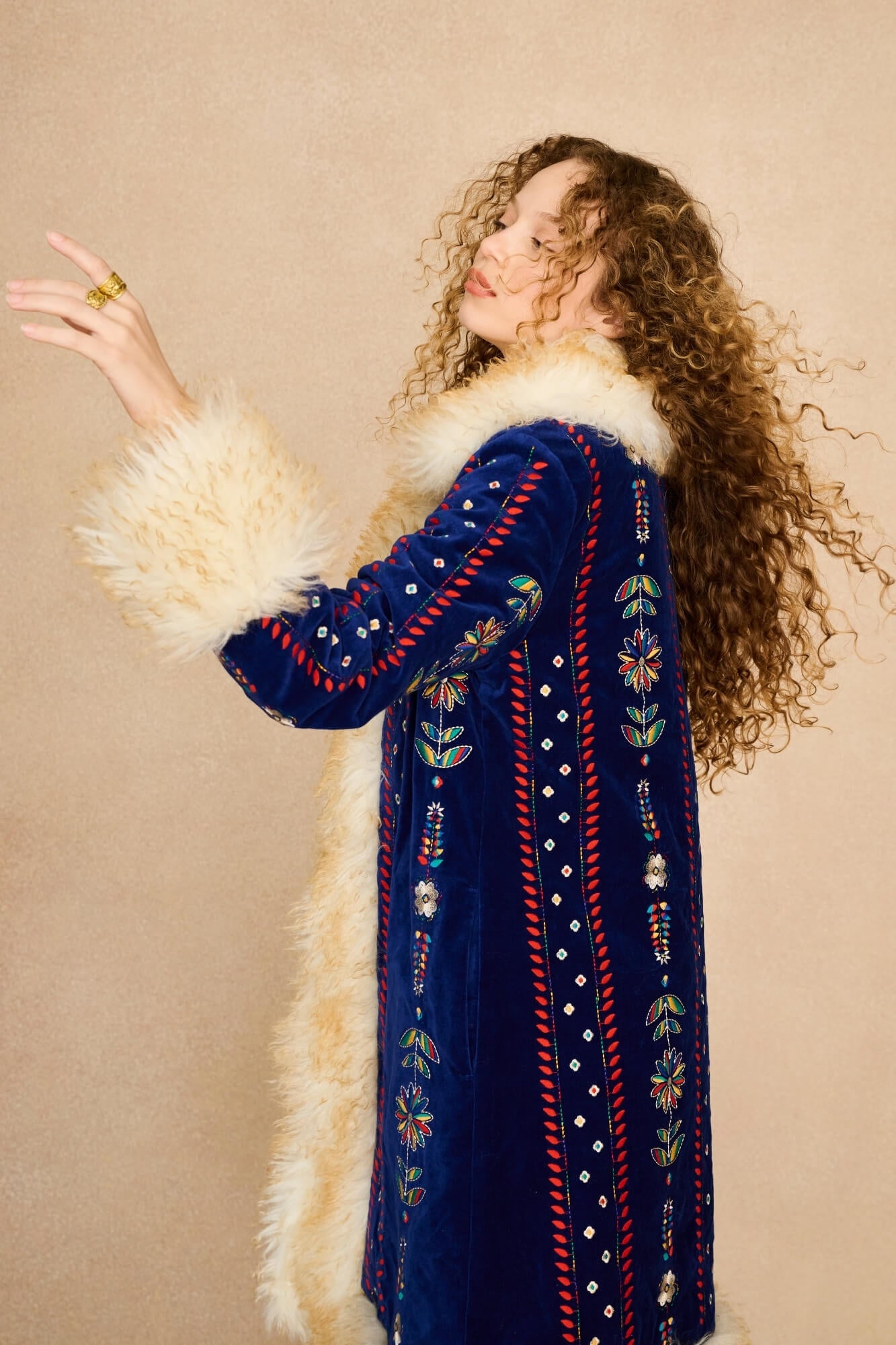 A woman with long curly brown hair models a long navy blue velvet Penny Lane coat with white fur trim and colourful embroidered details, posed against a solid beige background.