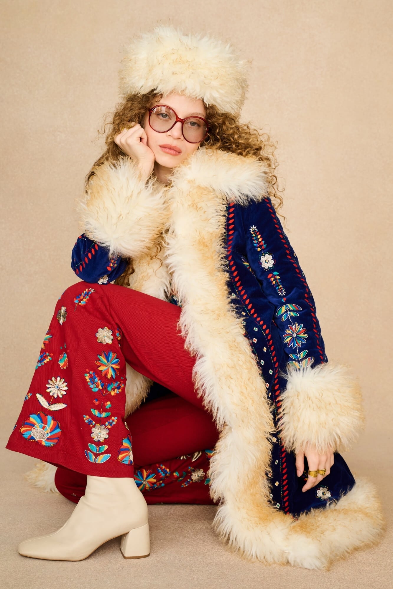 A model wearing a vintage-style folk-style outfit, featuring a navy blue embroidered Penny Lane coat with thick cream fur trim, red embroidered pants, a matching cream fur hat, and cream heeled boots, posed against a solid beige background.