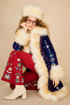 A model wearing a vintage-style folk-style outfit, featuring a navy blue embroidered Penny Lane coat with thick cream fur trim, red embroidered pants, a matching cream fur hat, and cream heeled boots, posed against a solid beige background.