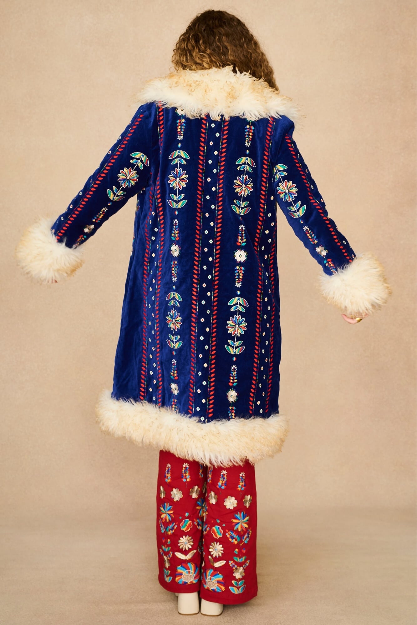 A model shown from the back wearing a long blue velvet Penny Lane coat with white faux-fur trim and colourful embroidered vertical patterns, paired with red patterned pants, against a plain beige studio background.
