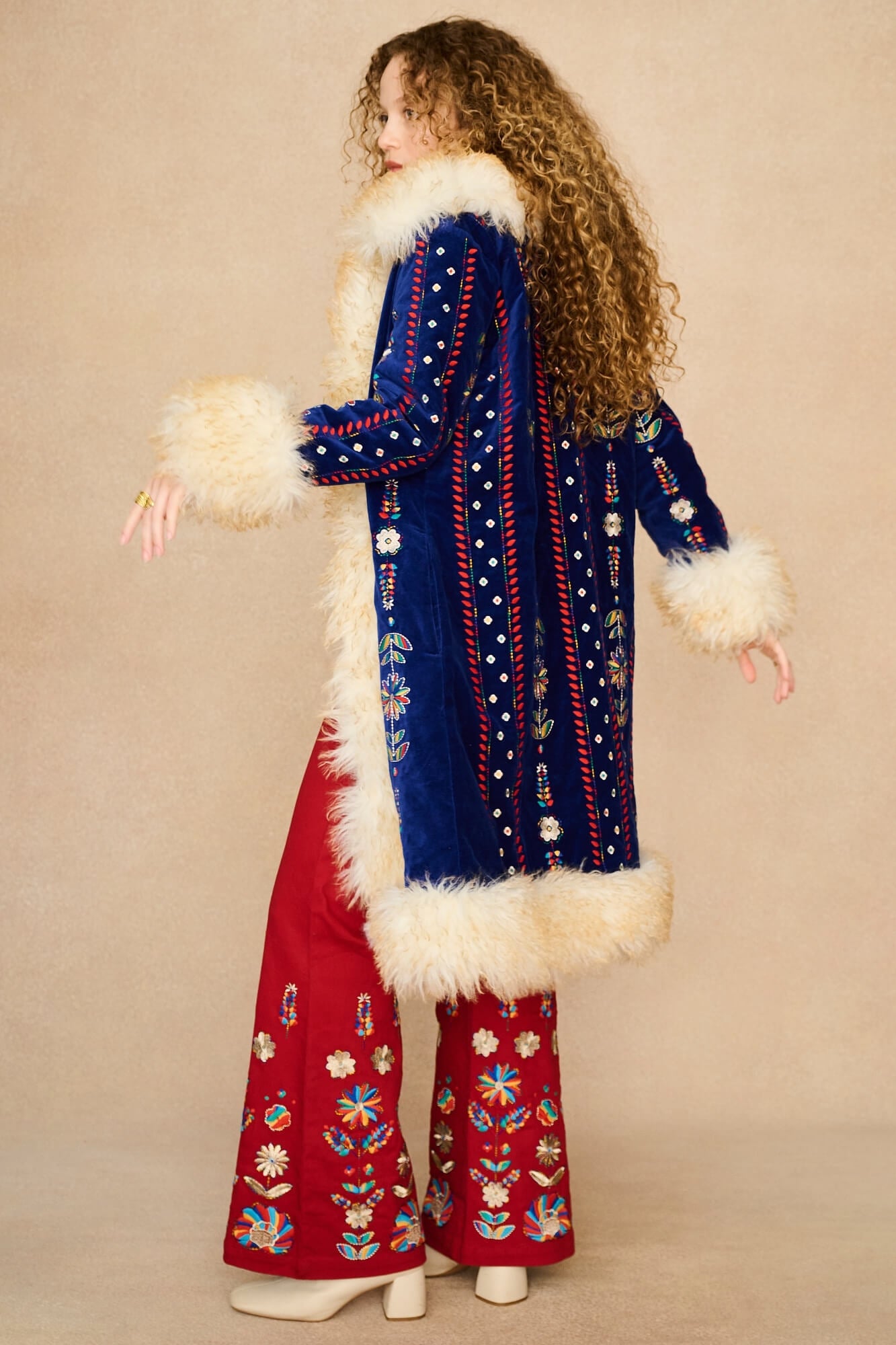 A woman stands in profile wearing a long blue velvet Penny lane coat by The Hippie Shake with cream fur trim and red wide-leg pants with floral embroidery, against a plain beige studio background.