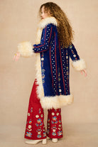 A woman stands in profile wearing a long blue velvet Penny lane coat by The Hippie Shake with cream fur trim and red wide-leg pants with floral embroidery, against a plain beige studio background.