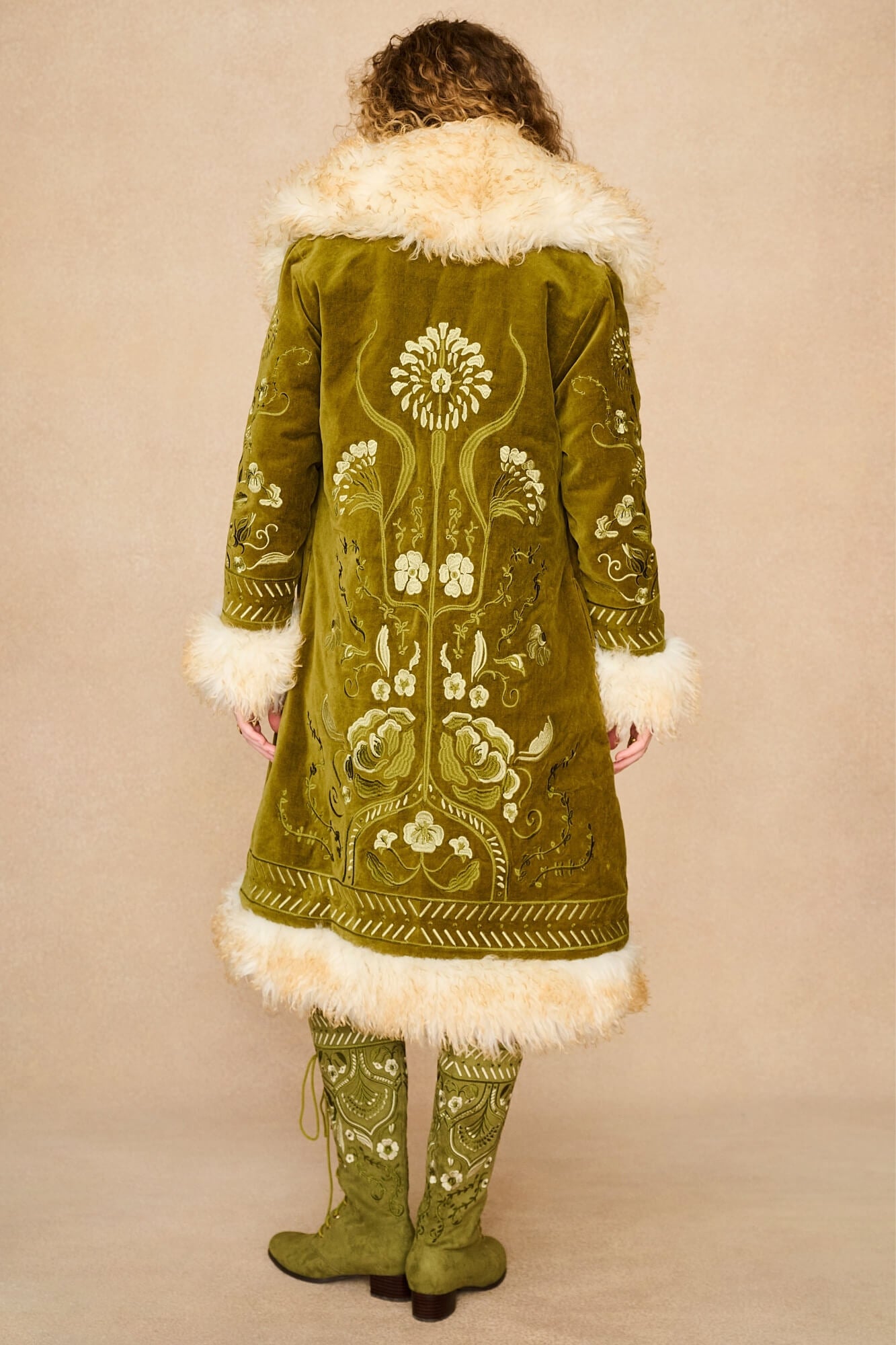 Model shown from the back wearing a long olive green embroidered Penny lane coat by The Hippie Shake with cream fur trim and matching embroidered green boots, against a plain beige studio background.
