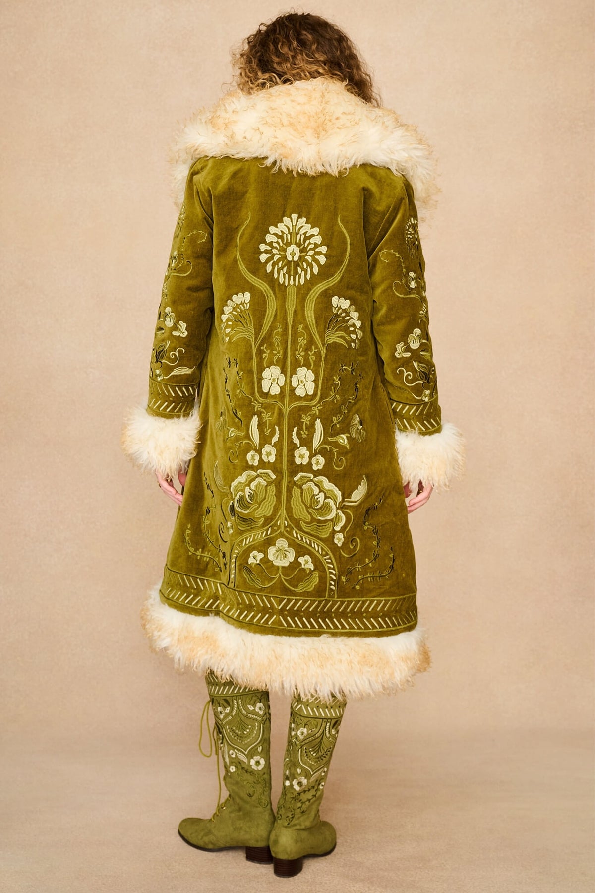 Model shown from the back wearing a long olive green embroidered Penny lane coat by The Hippie Shake with cream fur trim and matching embroidered green boots, against a plain beige studio background.