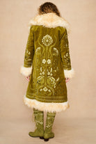 Model shown from the back wearing a long olive green embroidered Penny lane coat by The Hippie Shake with cream fur trim and matching embroidered green boots, against a plain beige studio background.