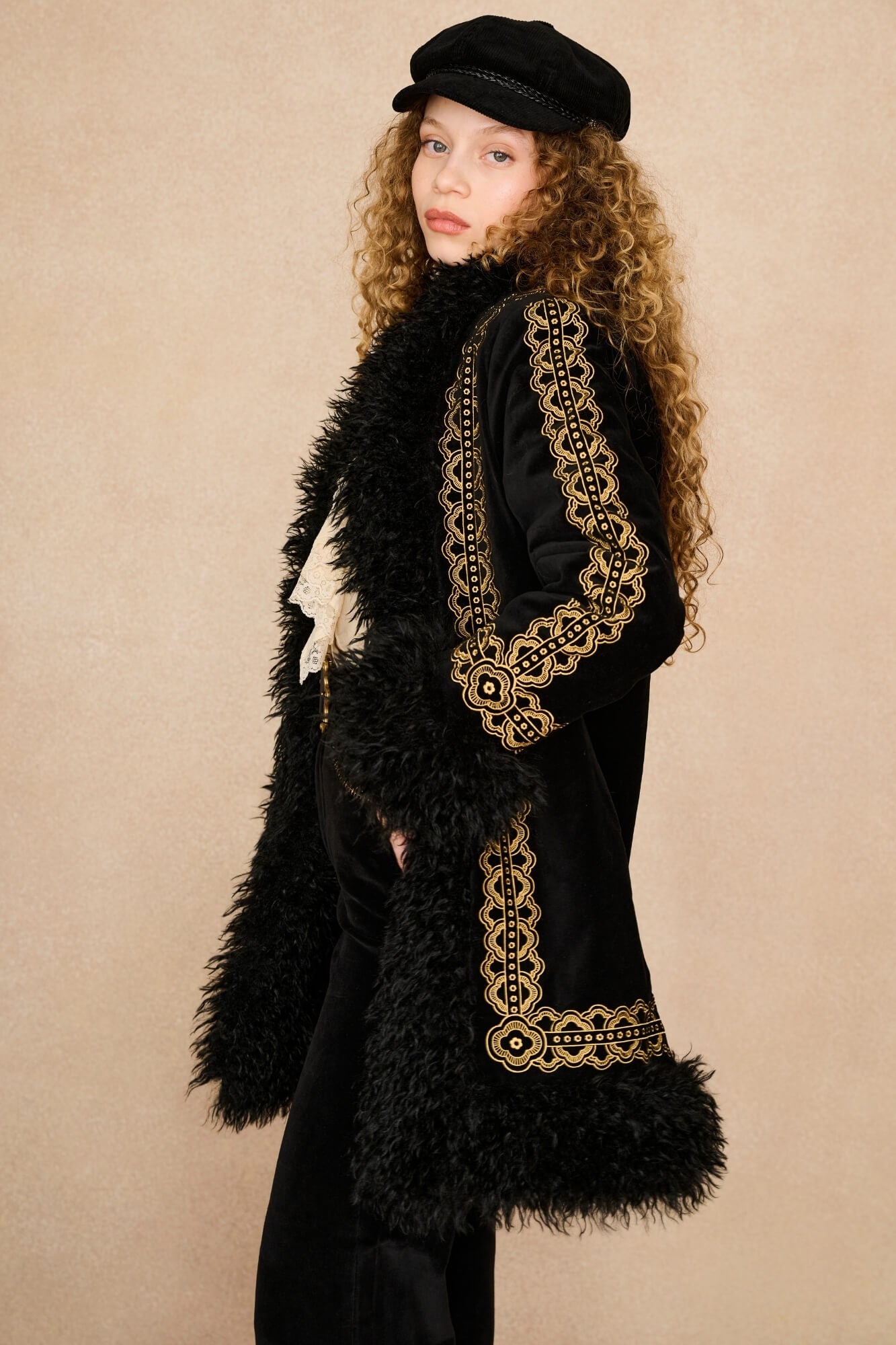 A model with long curly hair wearing a black velvet Penny lane coat by The Hippie Shake with ornate gold embroidery, a black faux-fur collar, a black beret, and black trousers, posed against a textured beige studio backdrop.