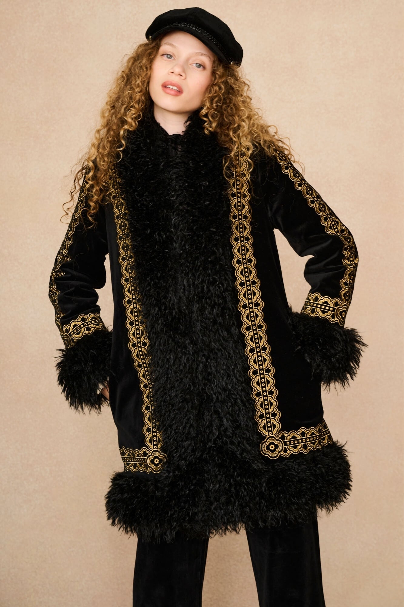 A model with long curly blonde hair wearing a black velvet and faux fur Penny Lane coat by The Hippie Shake with ornate gold embroidered borders, a black beret, and black pants, posed against a solid beige studio background.