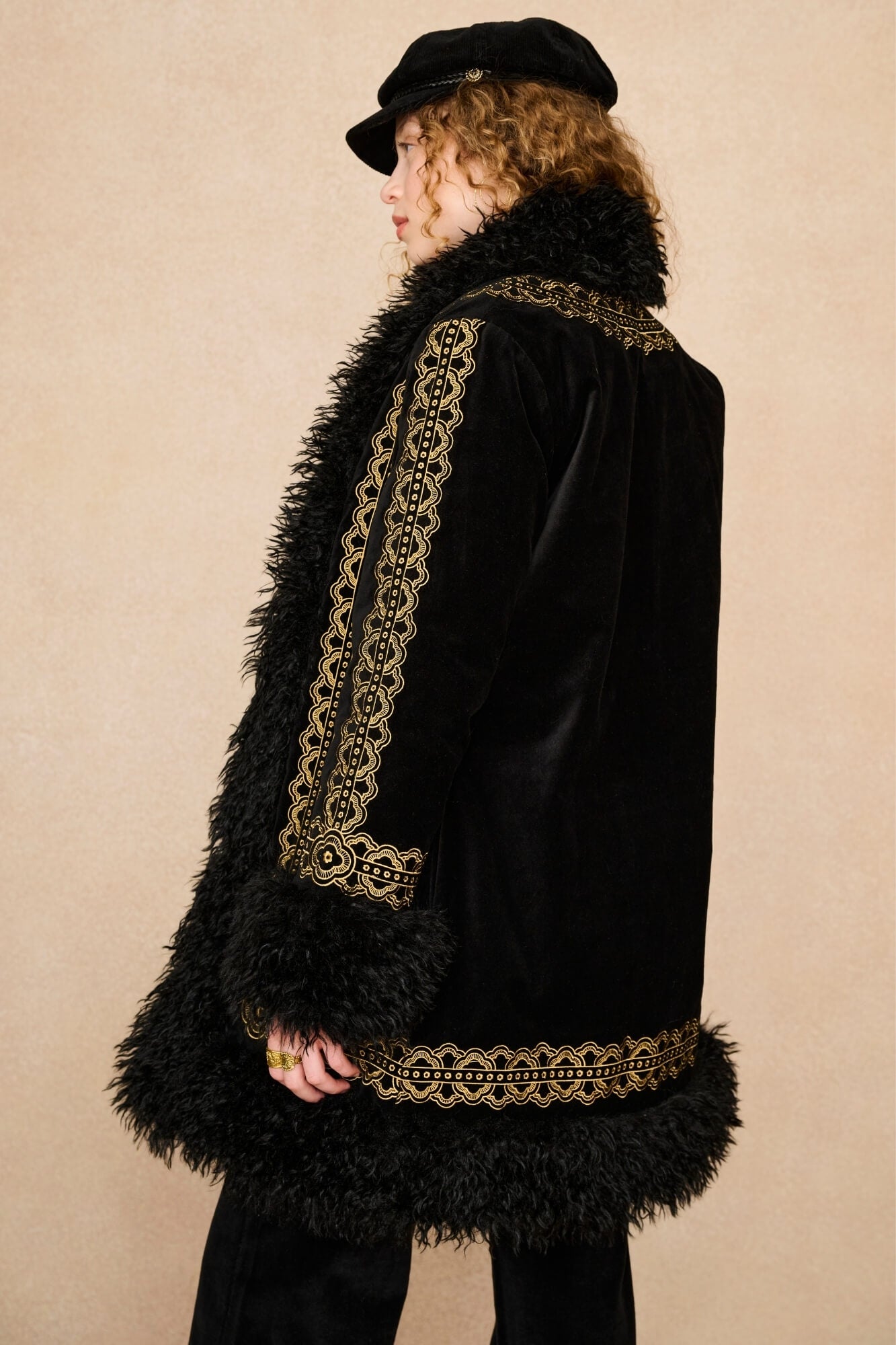 A person shown in side profile wearing a black velvet coat with ornate gold embroidered trim and a black feathered collar, along with a black cap, against a plain beige studio background.