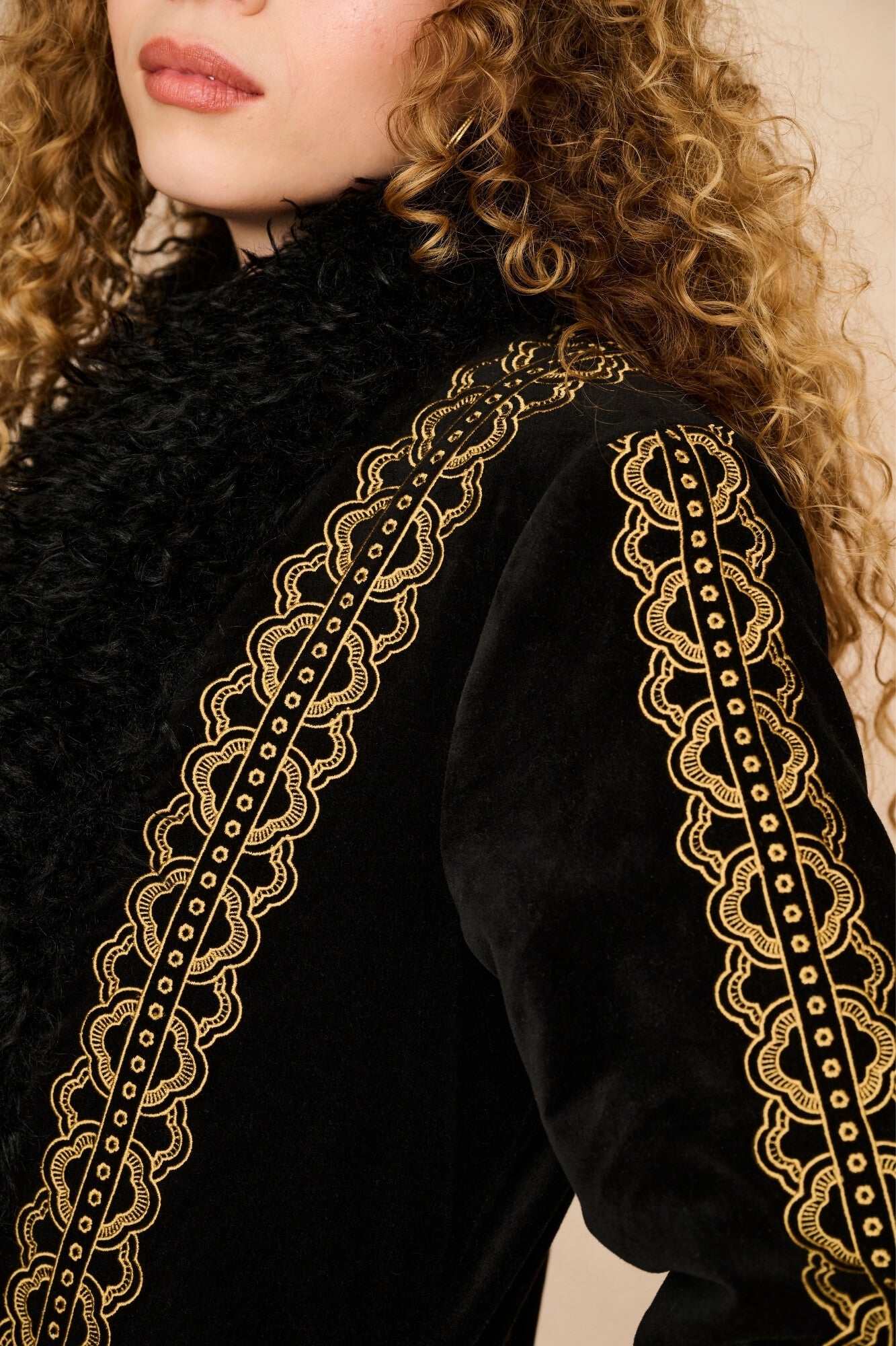 Close-up of a model wearing a black velvet jacket with intricate gold embroidered lace trim along the shoulders and collar.