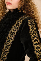 Close-up of a model wearing a black velvet jacket with intricate gold embroidered lace trim along the shoulders and collar.