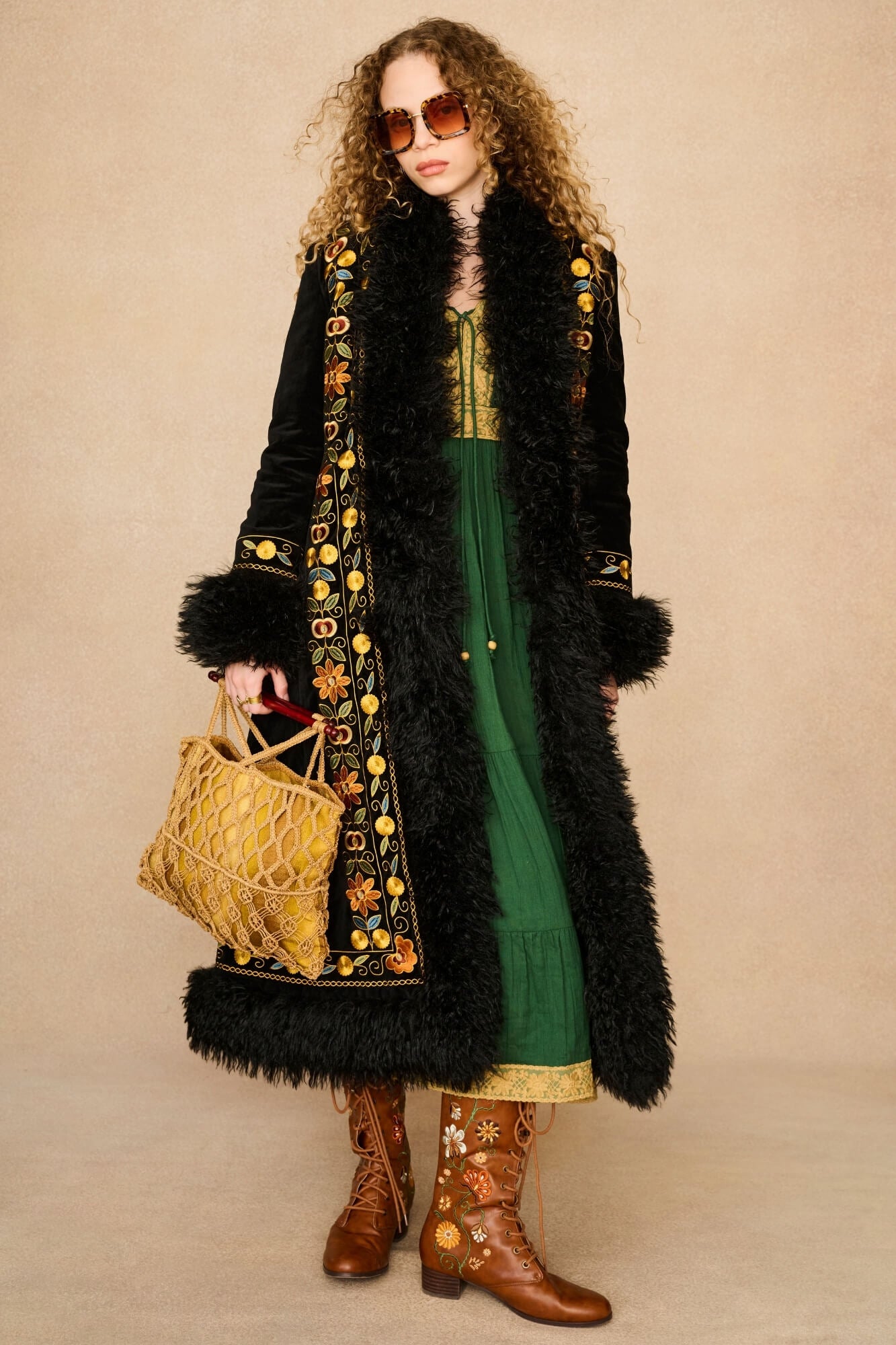 A woman stands against a plain beige studio background wearing a long black faux-fur coat with ornate gold floral embroidery, a green dress, brown western boots, and holding a woven tan tote bag.