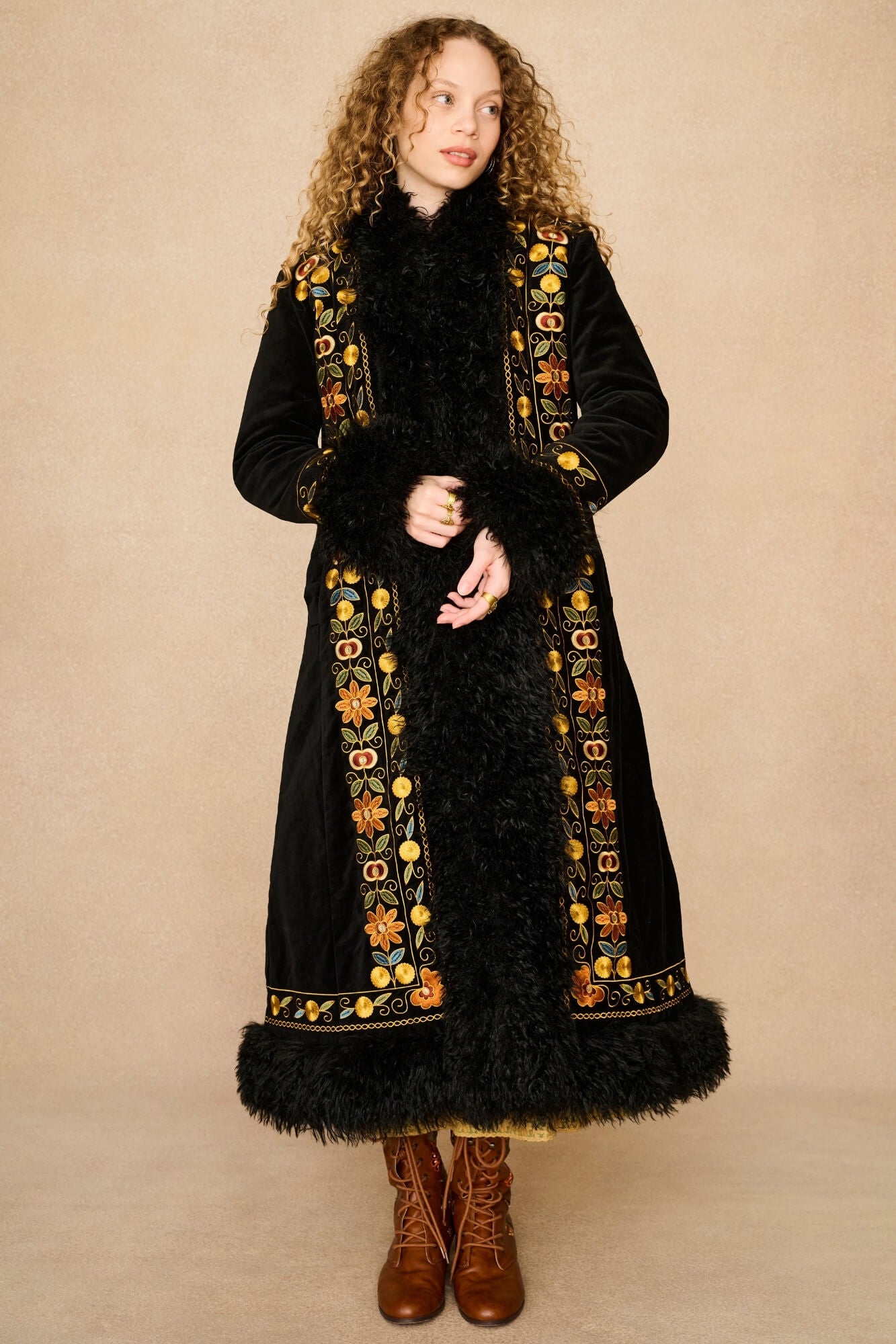 Model wearing a long black velvet Penny Lane coat by The Hippie Shake with black fur trim and ornate gold, orange, and yellow embroidered borders, paired with brown lace-up boots, standing against a plain beige studio backdrop.