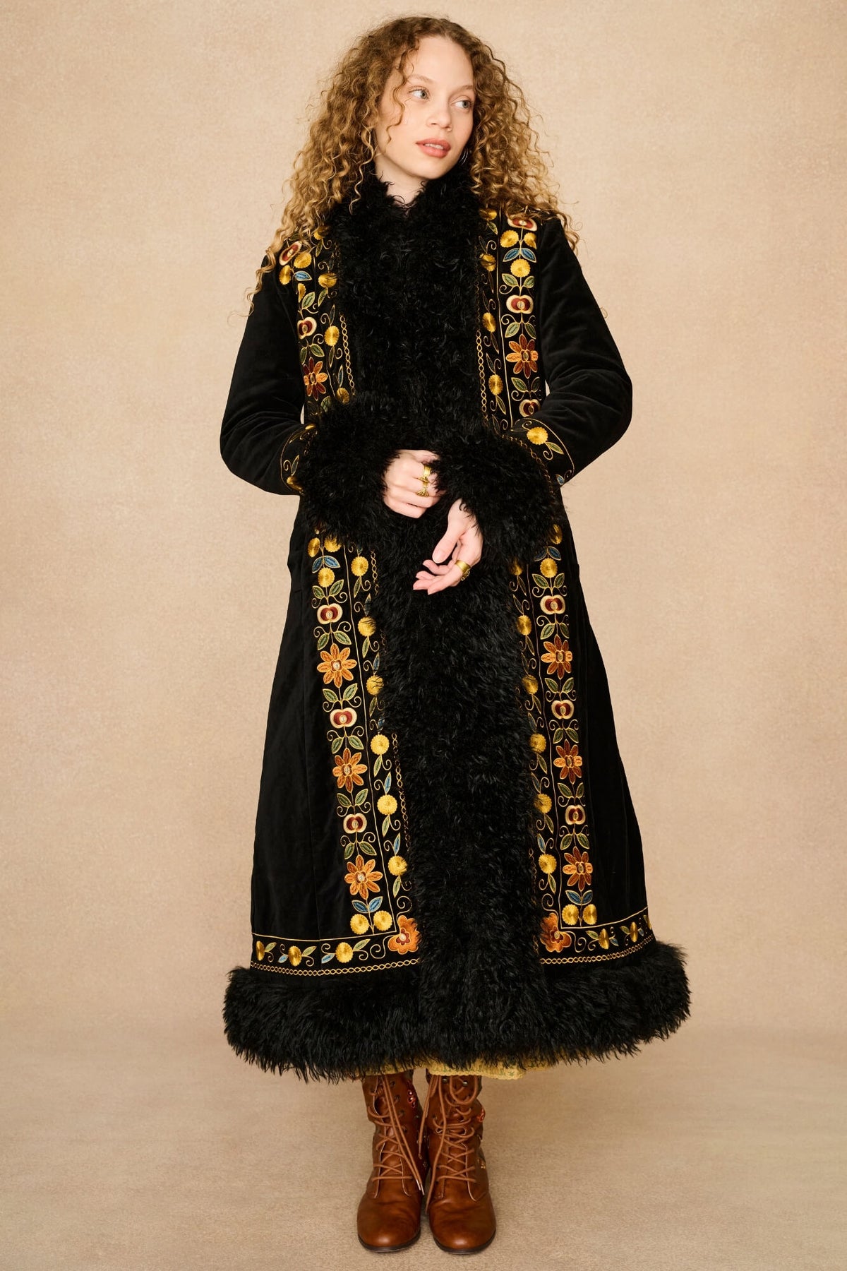 Model wearing a long black velvet Penny Lane coat by The Hippie Shake with black fur trim and ornate gold, orange, and yellow embroidered borders, paired with brown lace-up boots, standing against a plain beige studio backdrop.