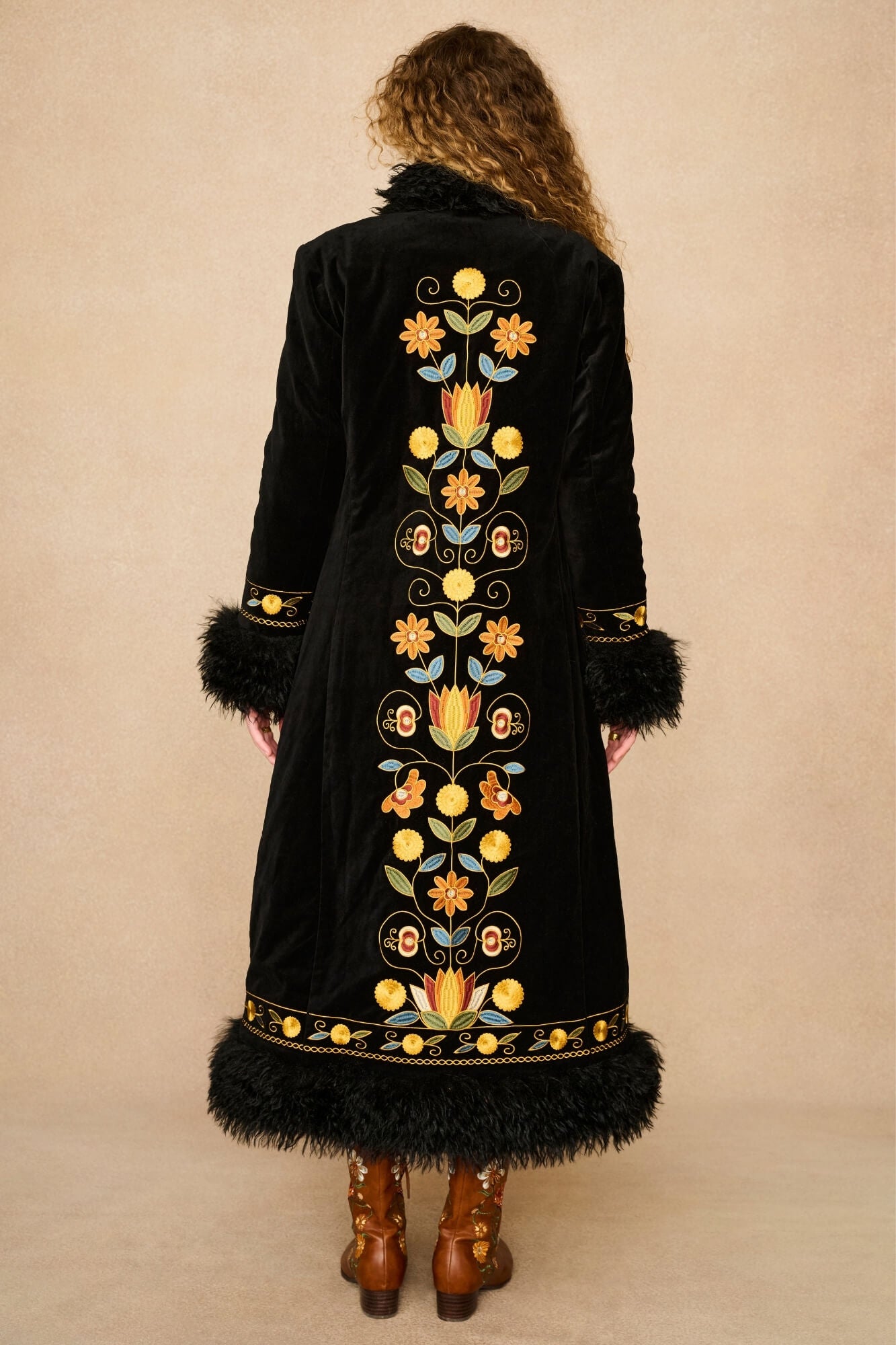 A woman viewed from behind wearing a long black velvet Penny lane coat by The Hippie Shake with colourful floral embroidery down the center, black feather trim at the collar and hem, and brown leather boots, against a plain beige studio background.