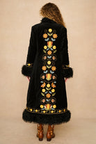 A woman viewed from behind wearing a long black velvet Penny lane coat by The Hippie Shake with colourful floral embroidery down the center, black feather trim at the collar and hem, and brown leather boots, against a plain beige studio background.