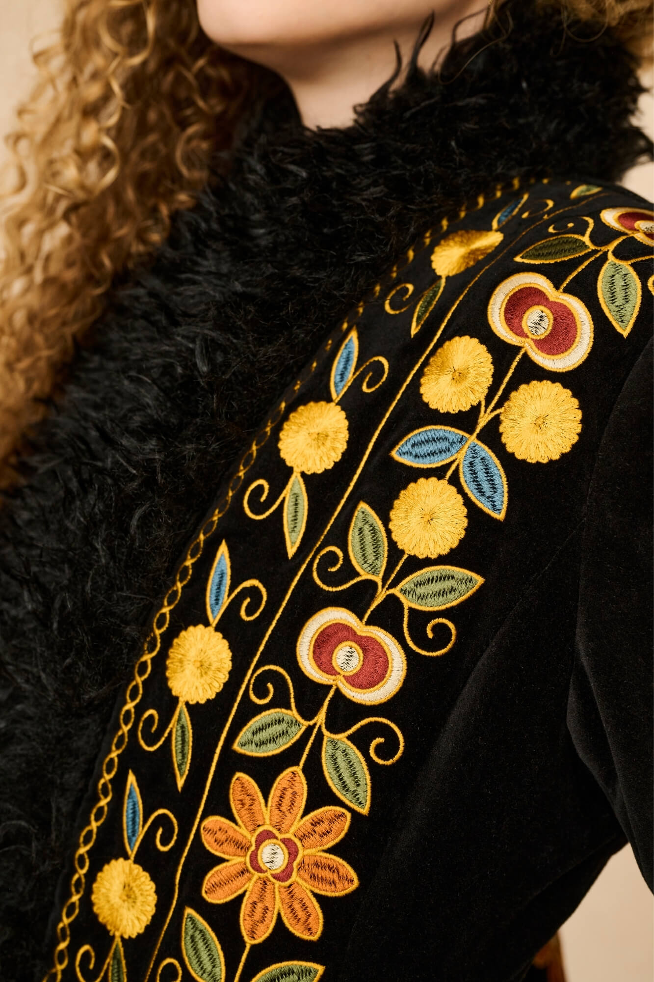 Close-up of a black velvet coat with a black faux-fur collar and ornate multicolor floral embroidery in yellow, gold, blue, and red along the neckline.